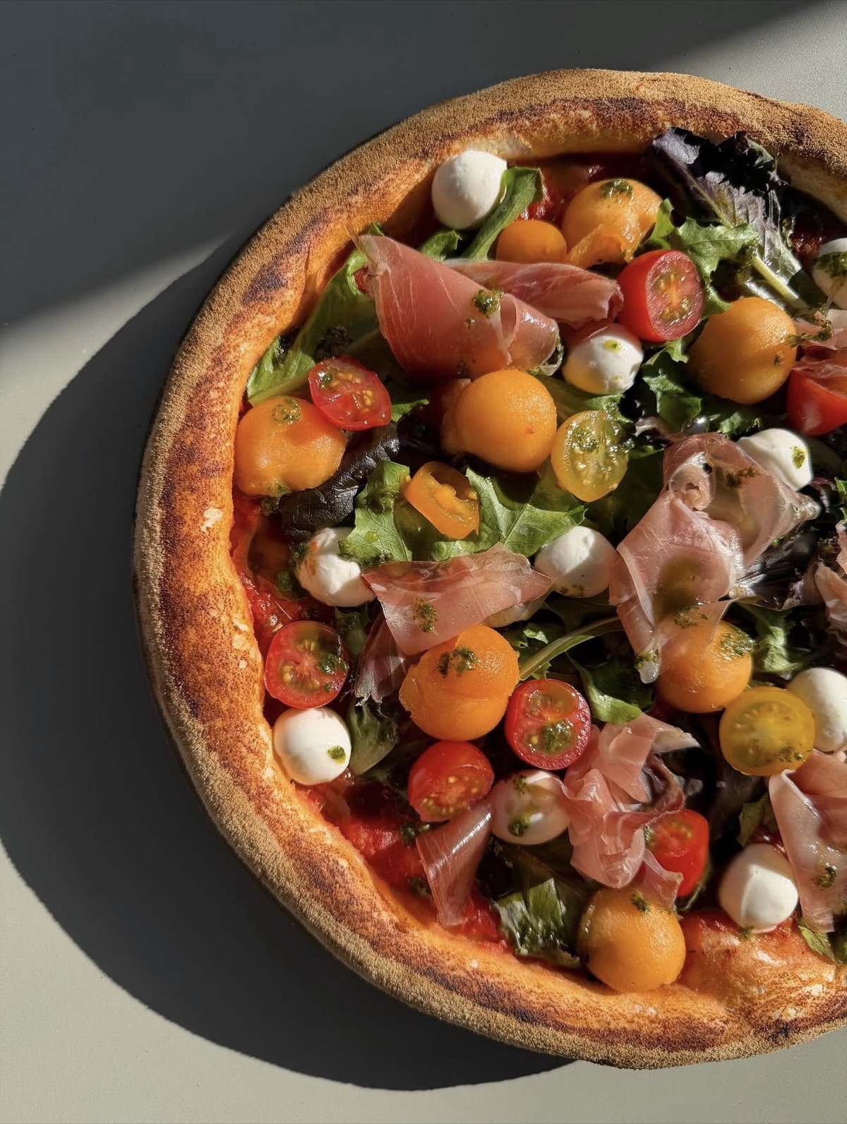 Pizza garnie de billes de melon, tomates cerises, mesclun, mozzarella et jambon cru